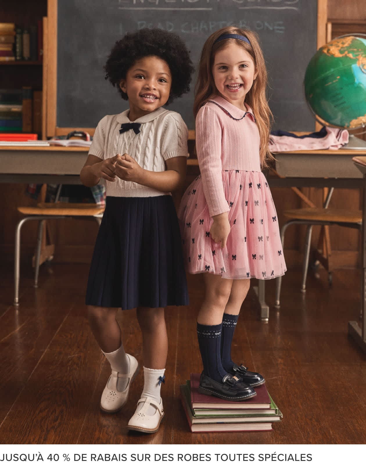 Deux jeunes filles, l'une aux cheveux bouclés, l'autre aux cheveux lisses, se tiennent dans une salle de classe, avec un tableau noir et des étagères en arrière-plan. Elles portent des robes et semblent sourire.
