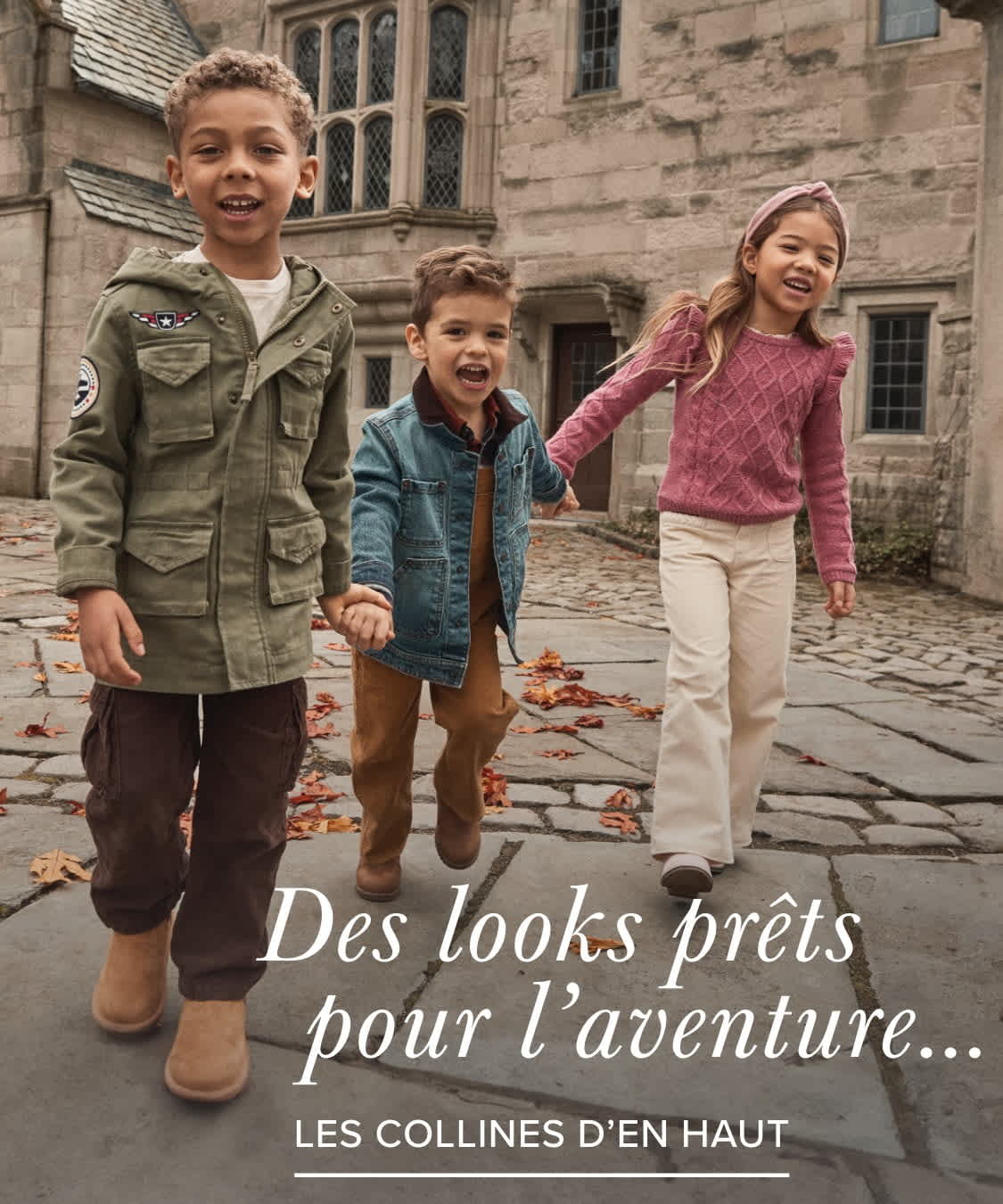 Three young children, a boy and two girls, are standing in front of a historic-looking building with stone walls and arched windows. The children are dressed in casual, adventure-ready outfits, suggesting an outdoor or exploratory theme.