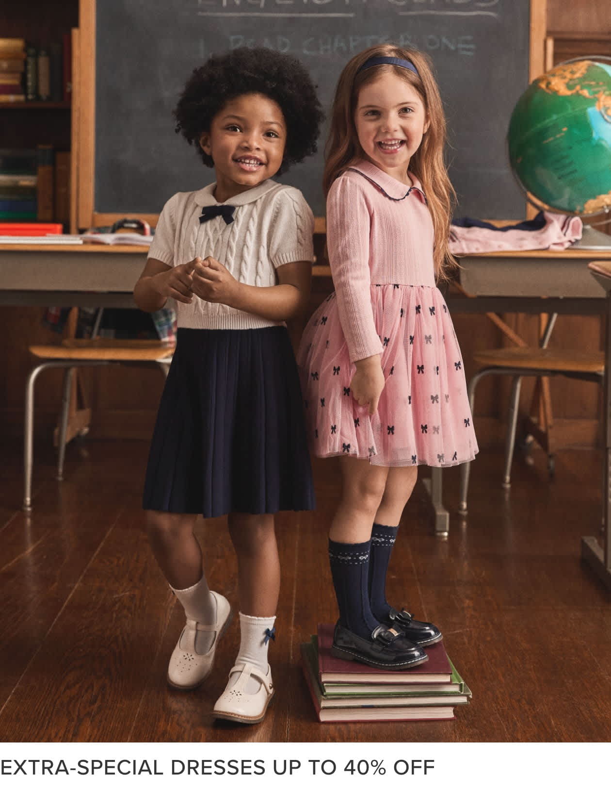 Two young girls, one with curly hair and the other with straight hair, are standing in a classroom setting with a chalkboard and bookshelves visible in the background. The girls are wearing dresses and appear to be smiling.