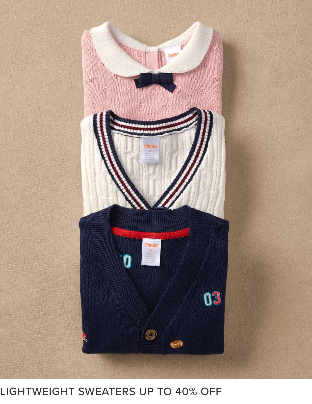 The image shows a stack of neatly folded children's sweaters in various colors and patterns, including a pink sweater with a bow, a striped sweater, and a navy blue cardigan with a patch. The sweaters are displayed against a beige background, likely in a retail setting.