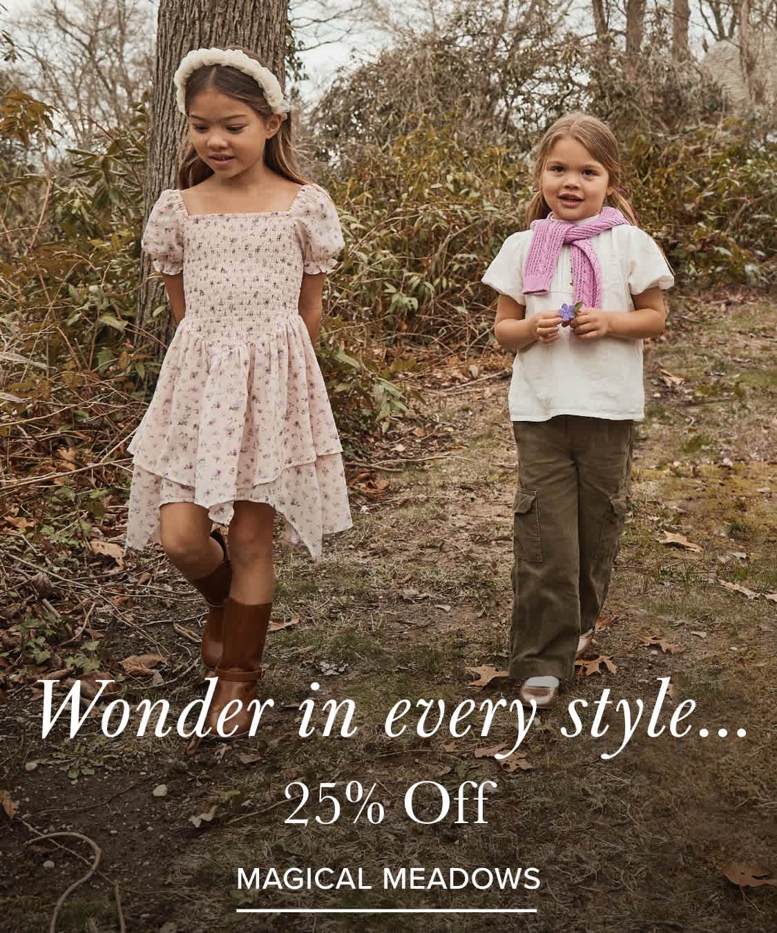 Two young girls, one wearing a pink floral dress and the other a white top, stand in a wooded area with trees and foliage in the background.