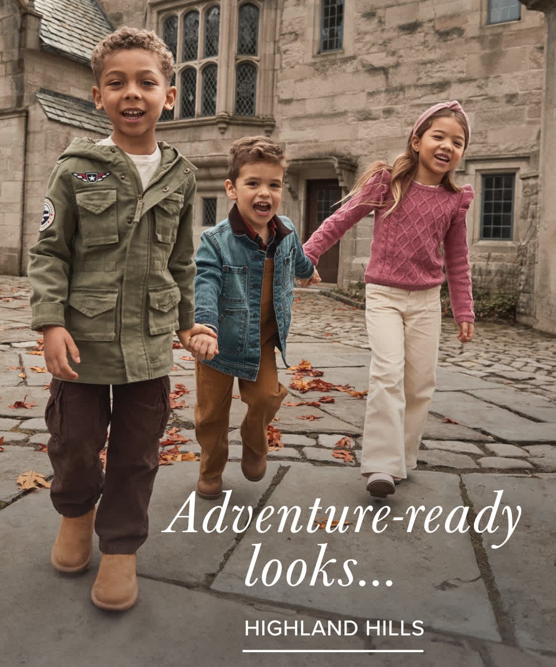 Three young children, a boy and two girls, are standing in front of a historic-looking building with stone walls and arched windows. The children are dressed in casual, adventure-ready outfits, suggesting an outdoor or exploratory theme.