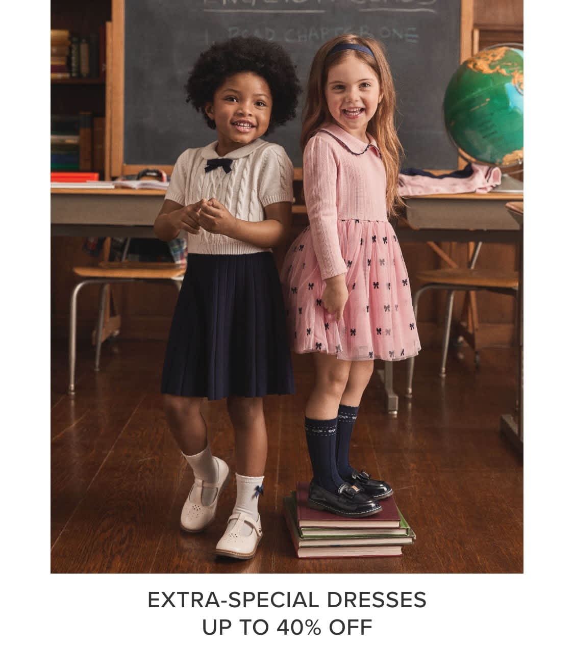 Two young girls, one with curly dark hair and the other with long blonde hair, are standing in a classroom setting with a chalkboard and other educational props in the background. The girls are wearing dresses, one in a pink pattern and the other in a solid navy color, and appear to be smiling and posing for the camera.