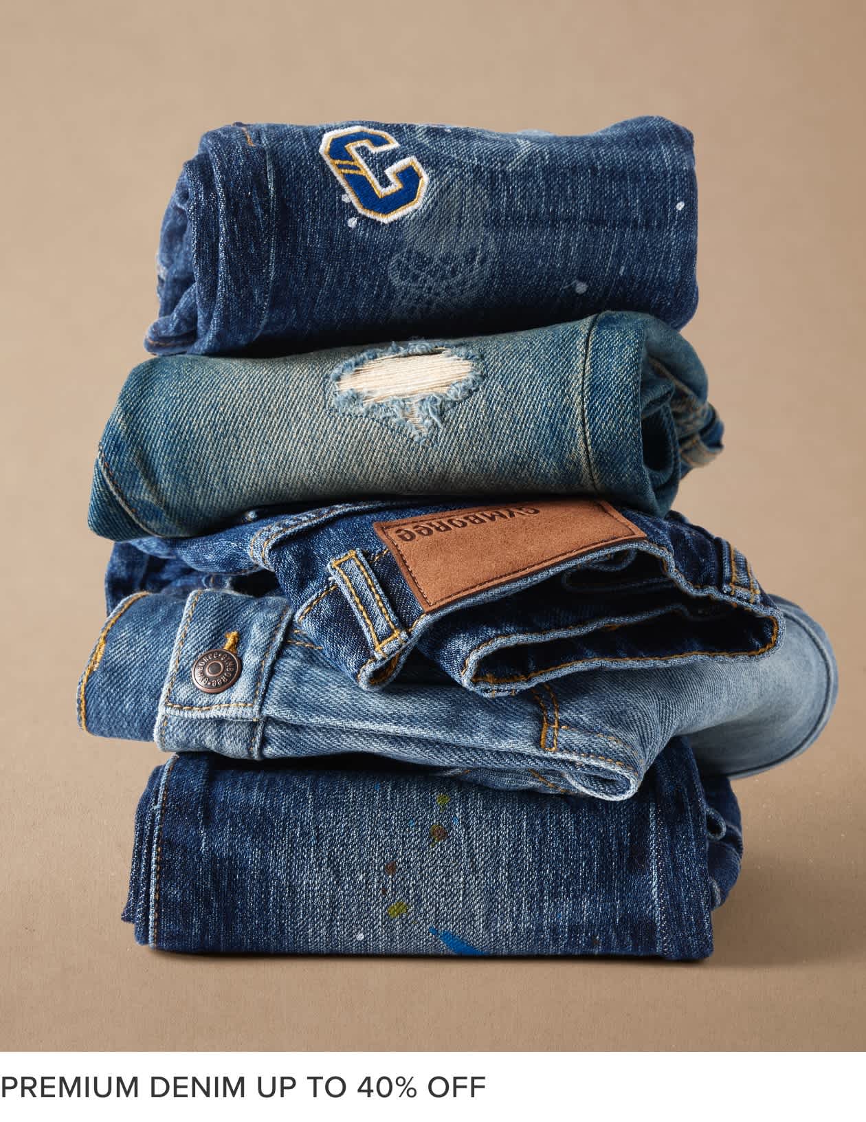A stack of various denim clothing items, including jeans, jackets, and a towel, against a beige background.