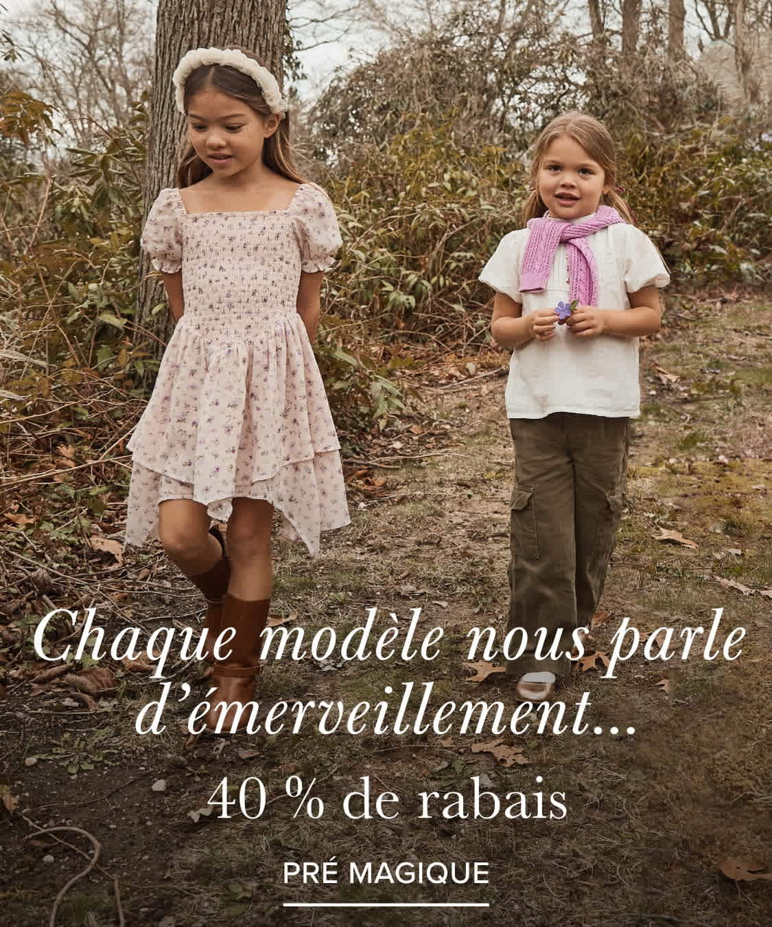 Two young girls, one wearing a pink floral dress and the other a white top, stand in a wooded area with trees and foliage in the background.