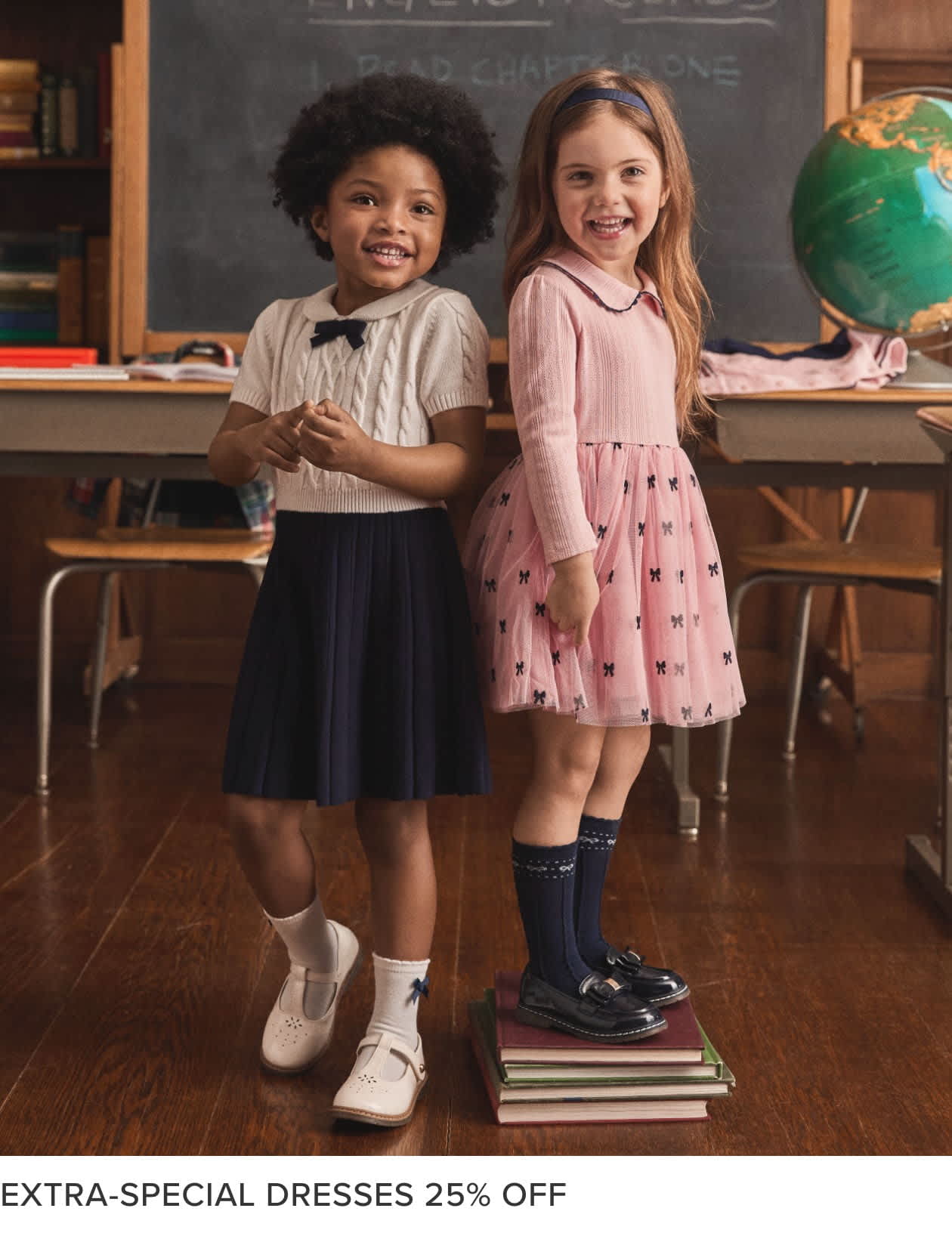 Two young girls, one with curly hair and the other with straight hair, are standing in a classroom setting with a chalkboard and bookshelves visible in the background. The girls are wearing dresses and appear to be smiling.