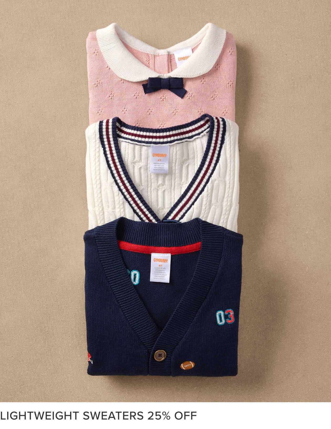 The image shows a stack of neatly folded children's sweaters in various colors and patterns, including a pink sweater with a bow, a striped sweater, and a navy blue cardigan with a patch. The sweaters are displayed against a beige background, likely in a retail setting.