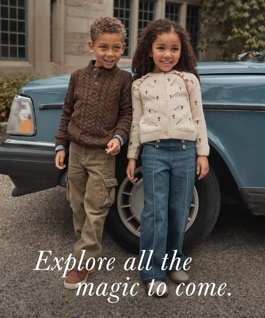 Two young people, a boy and a girl, stand together in front of an old building with large windows, with a vintage-style car in the background. The text overlay encourages the viewer to "Explore all the magic to come."