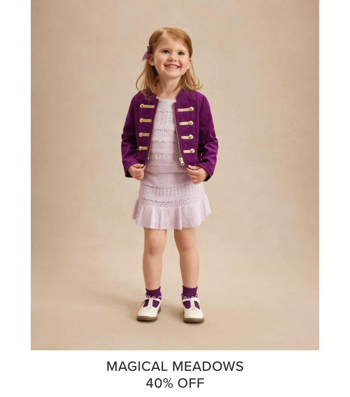 A young girl with blonde hair and a bright smile, wearing a purple jacket and a white dress, stands against a beige background.
