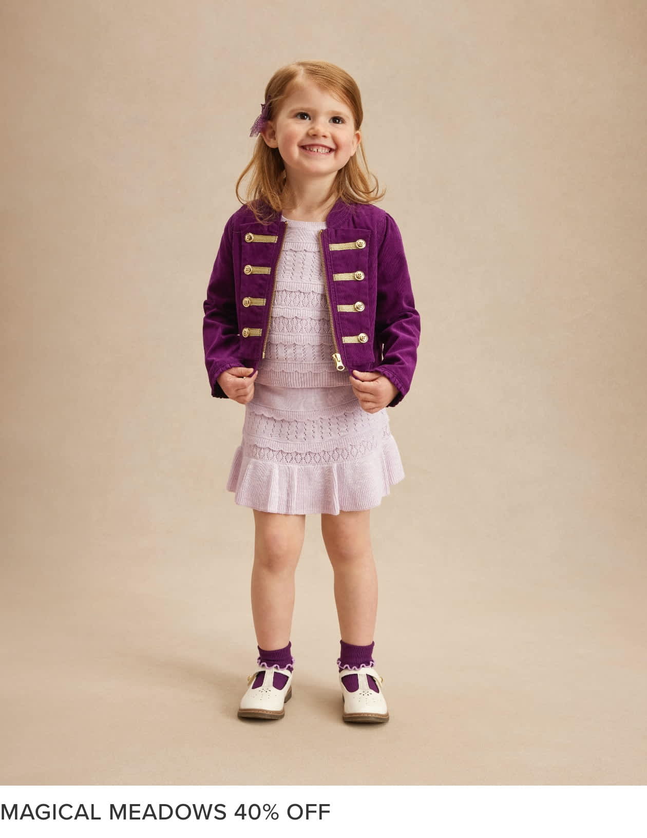 A young girl with blonde hair and a bright smile, wearing a purple jacket and a white dress, stands against a beige background.