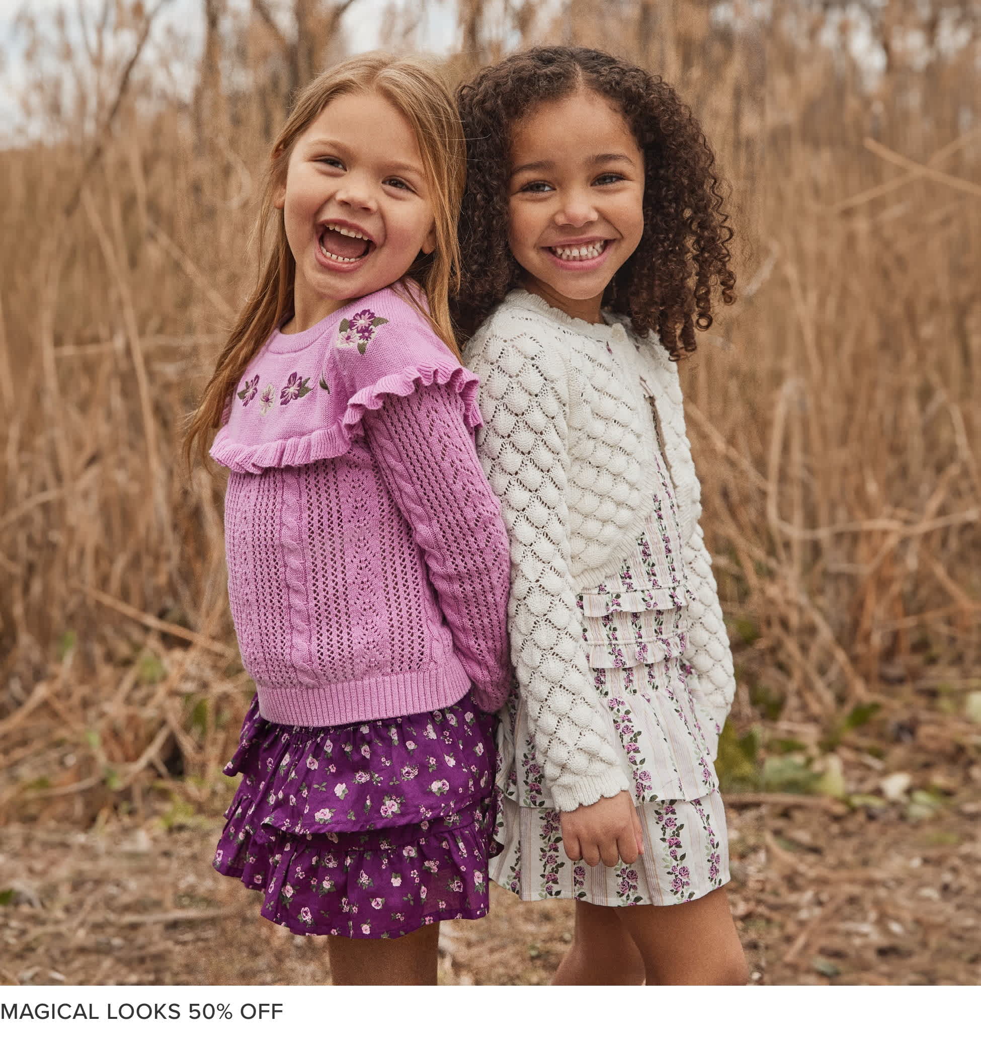 Two young girls, one with long blonde hair and the other with curly dark hair, are standing together and smiling brightly in a field of tall grass and bare trees in the background.