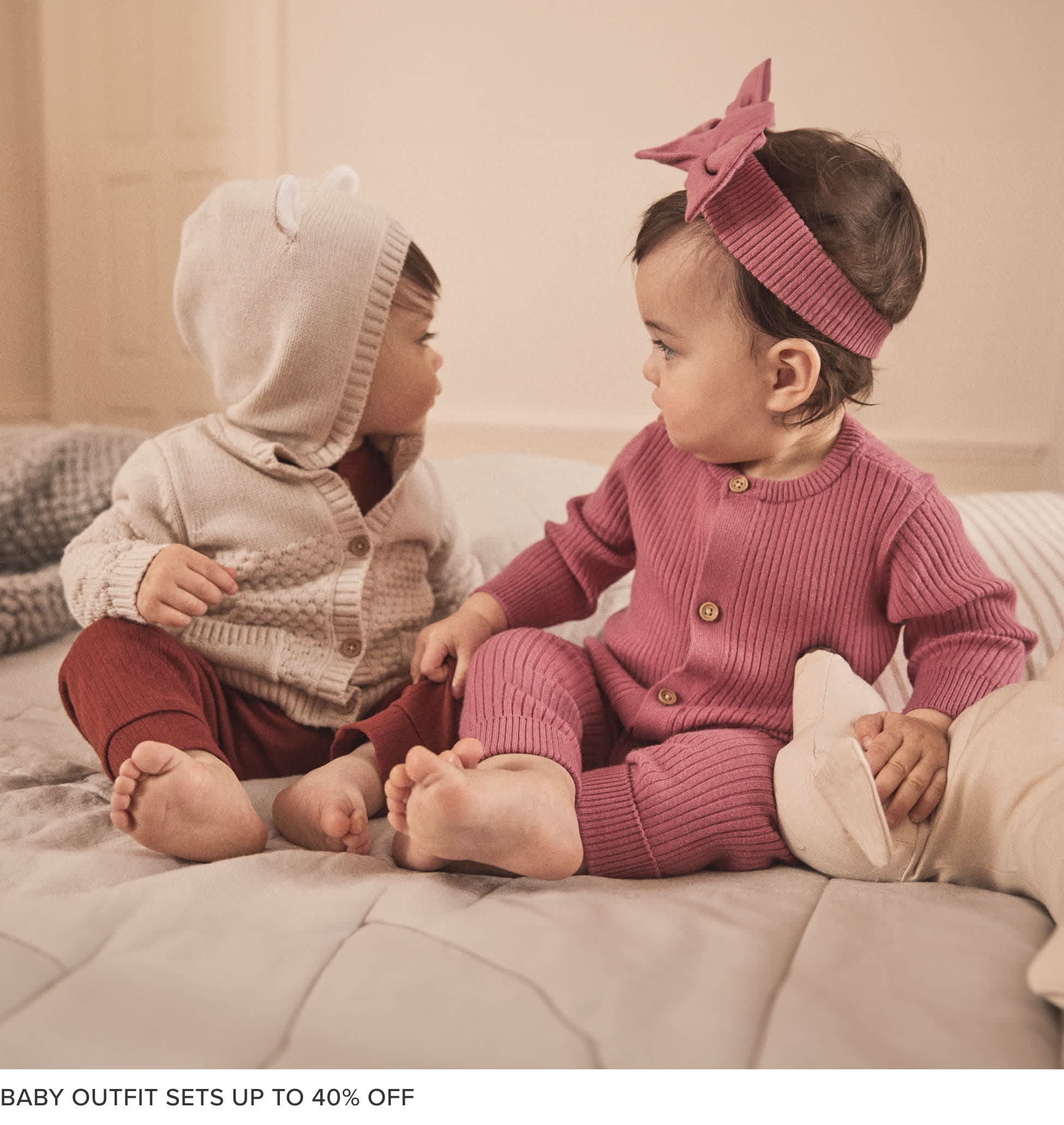 Two young children, a boy and a girl, sitting on a couch and wearing matching pink and gray knitted outfits. The background appears to be a warm, cozy living room setting.