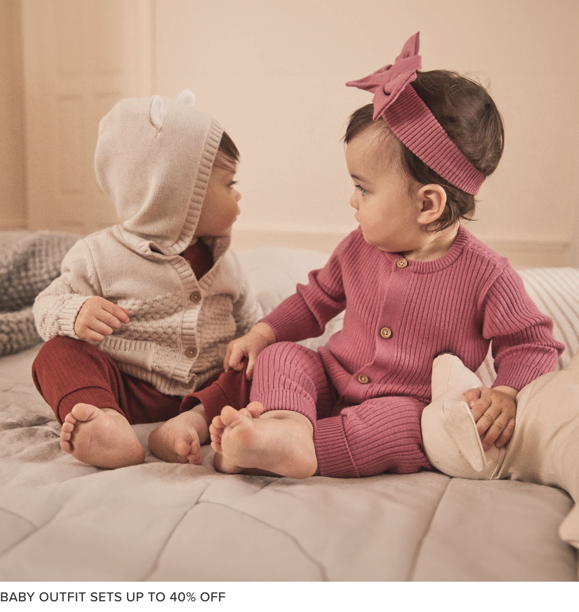 Two young children, a boy and a girl, sitting on a couch and wearing matching pink and gray knitted outfits. The background appears to be a warm, cozy living room setting.