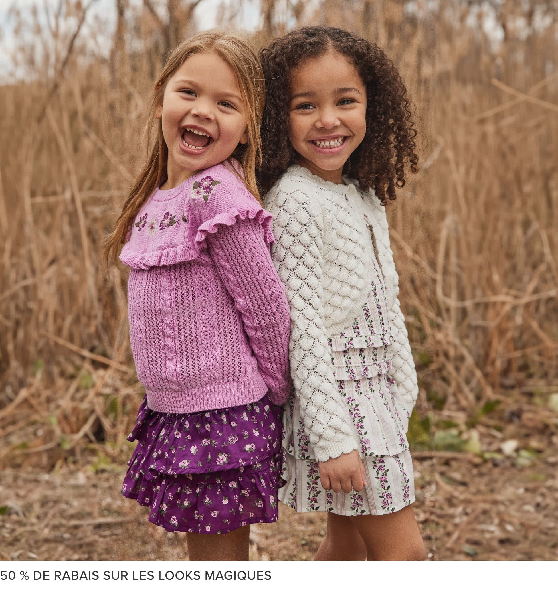 Deux jeunes filles, l'une aux longs cheveux blonds et l'autre aux cheveux noirs bouclés, se tiennent ensemble dans un champ d'herbes hautes, toutes deux vêtues de vêtements colorés et à motifs et souriant vivement.