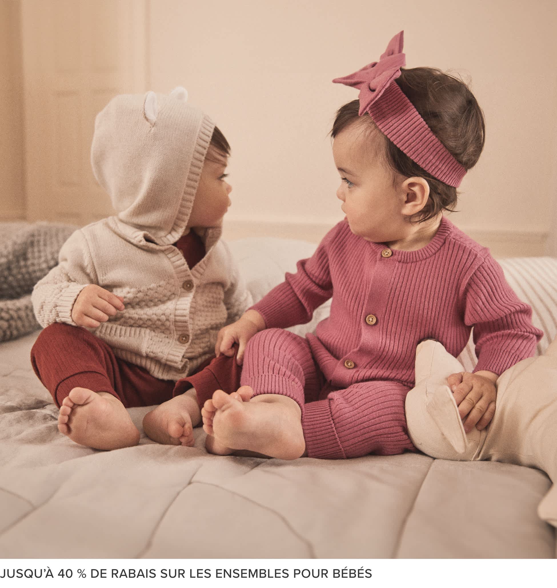 Deux jeunes enfants, un garçon et une fille, assis sur un canapé et portant des tenues tricotées assorties rose et grise. L'arrière-plan semble être un salon chaleureux et confortable.
