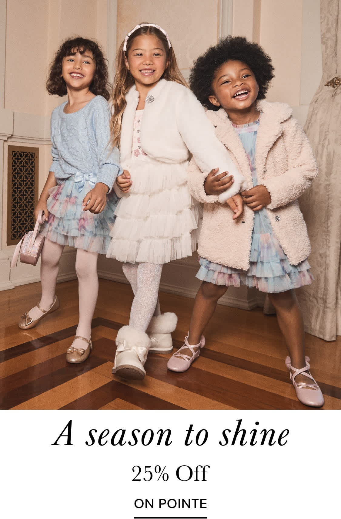 La imagen muestra a tres mujeres jóvenes vestidas con elegantes trajes blancos y de colores pastel con volantes, paradas juntas y sonriendo alegremente en lo que parece ser un gran y ornamentado entorno interior con cortinas y detalles arquitectónicos en el fondo.