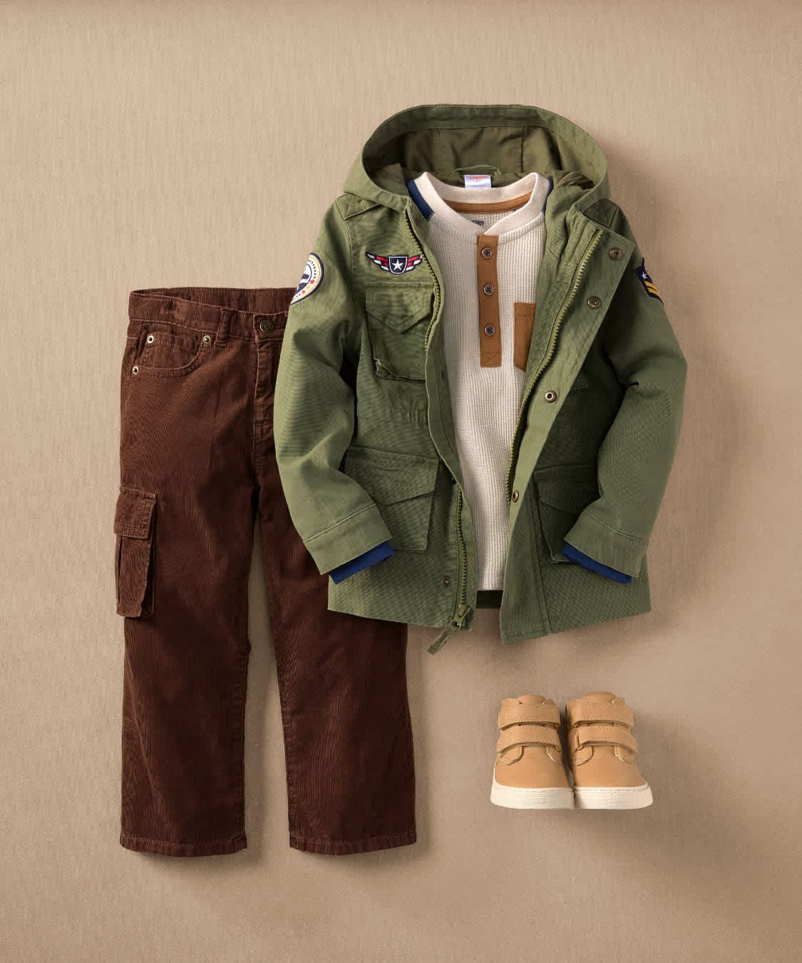 A collection of children's clothing items, including a green hooded jacket, brown pants, and beige shoes, arranged on a beige background.