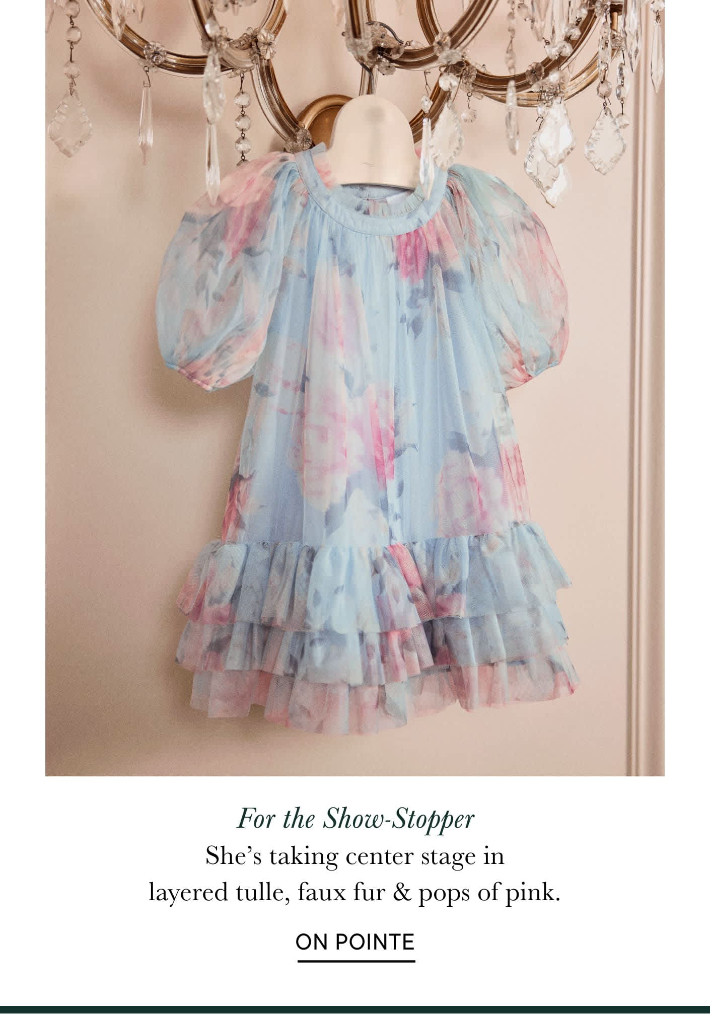 Un delicado vestido de tul en tonos pastel, con mangas abullonadas y capas con volantes, cuelga de una lámpara de araña, creando una composición caprichosa y femenina.