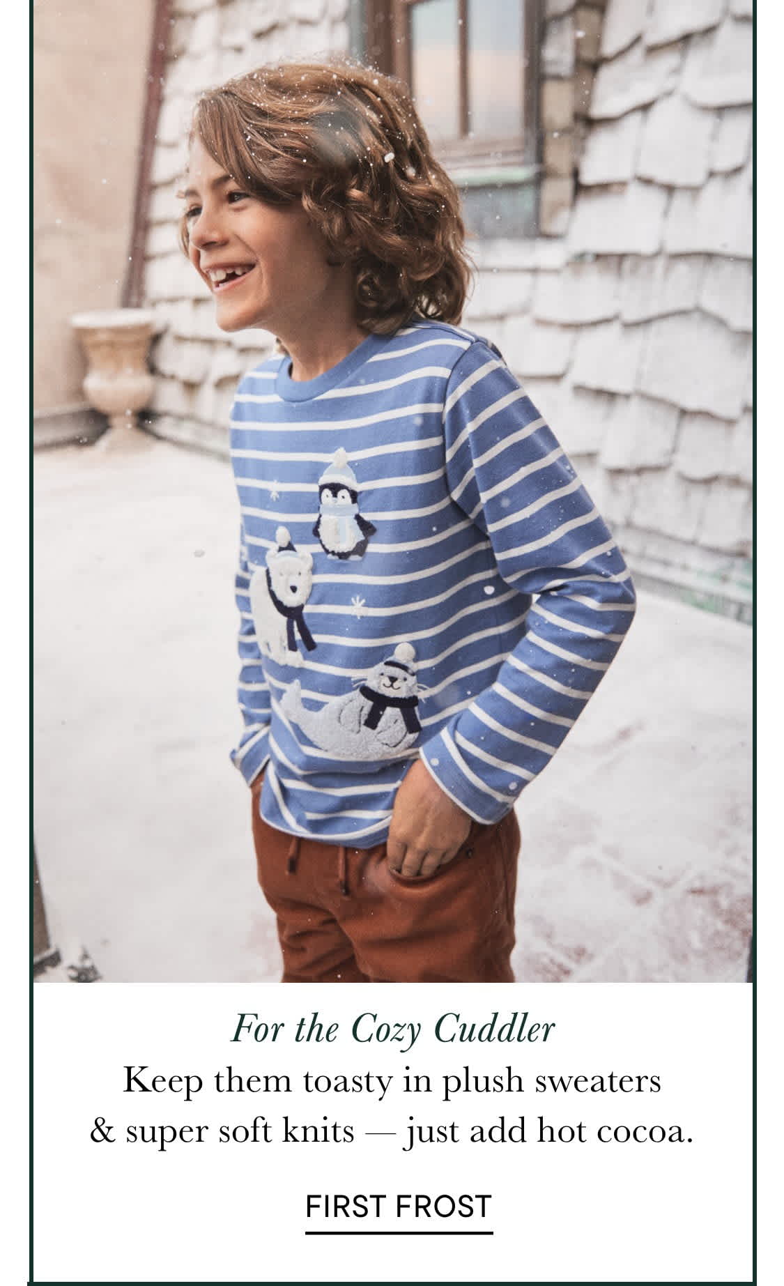 Una niña pequeña con el pelo rizado y un suéter a rayas está de pie frente a un edificio de madera nevado, sonriendo radiante.