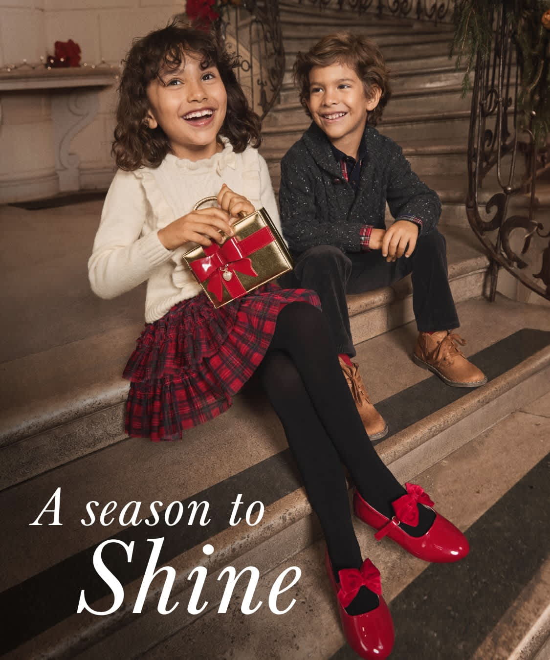 Two people, a man and a woman, are standing in a festive setting with holiday decorations in the background. They are dressed in stylish holiday attire, with the woman wearing a plaid skirt and the man in a dark jacket, creating a visually appealing scene.