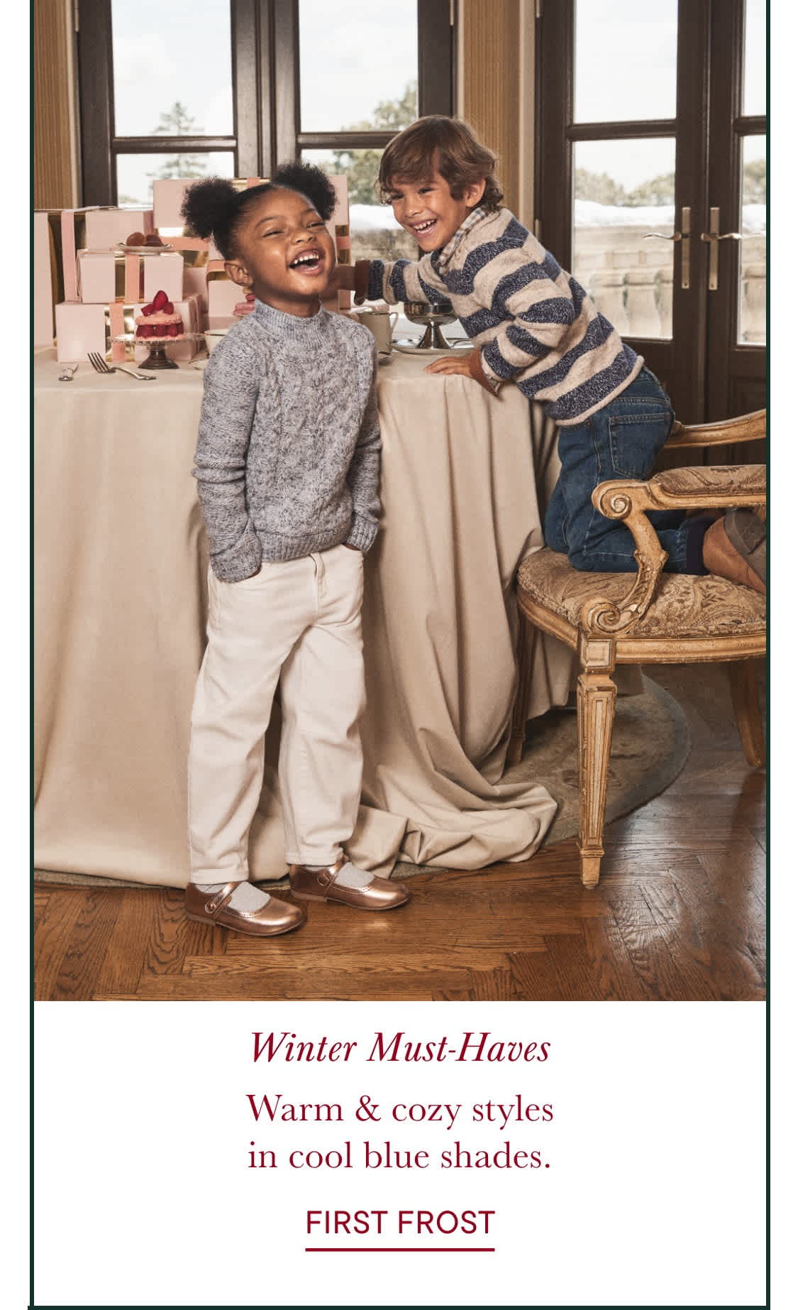 Two young children, a boy and a girl, are sitting on a couch in a cozy, warm-toned room with large windows overlooking a snowy outdoor scene. The children are smiling and appear to be enjoying each other's company.