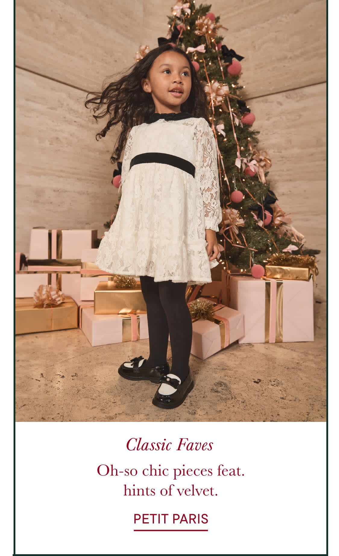 A young girl in a white lace dress stands in front of a Christmas tree, surrounded by wrapped gifts on a wooden floor.