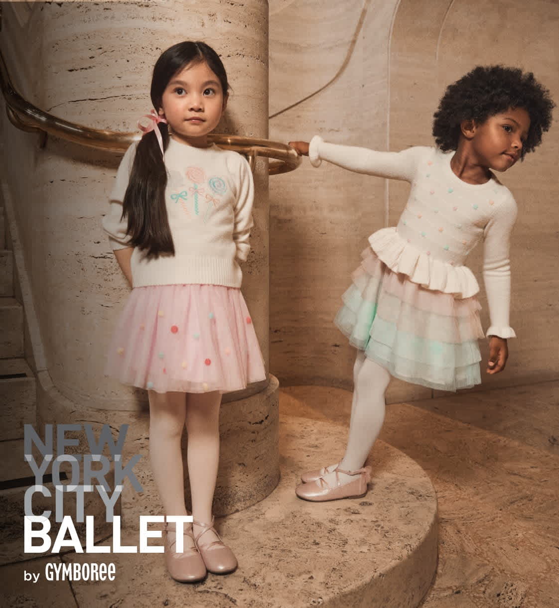 Two young girls, one with long dark hair and the other with curly hair, are dressed in ballet attire and posing in a studio setting with a textured wall backdrop.