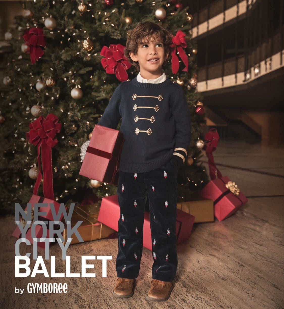 A young child wearing a navy blue sweater with gold buttons stands in front of a Christmas tree, holding a pink gift box, surrounded by additional wrapped presents.