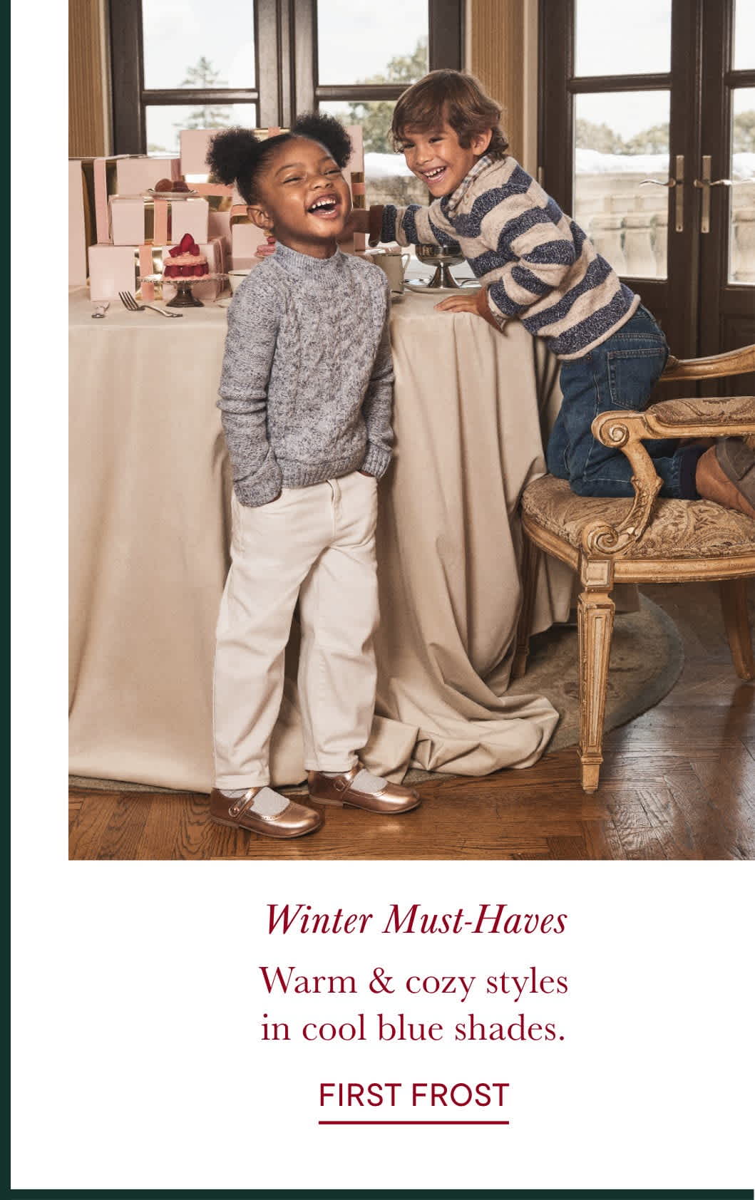 The image shows two young children, a boy and a girl, sitting on a couch in a cozy, warm-toned room. The children are dressed in winter clothing, with the boy wearing a striped sweater and the girl wearing a gray sweater. The background features a window, suggesting a comfortable, indoor setting.