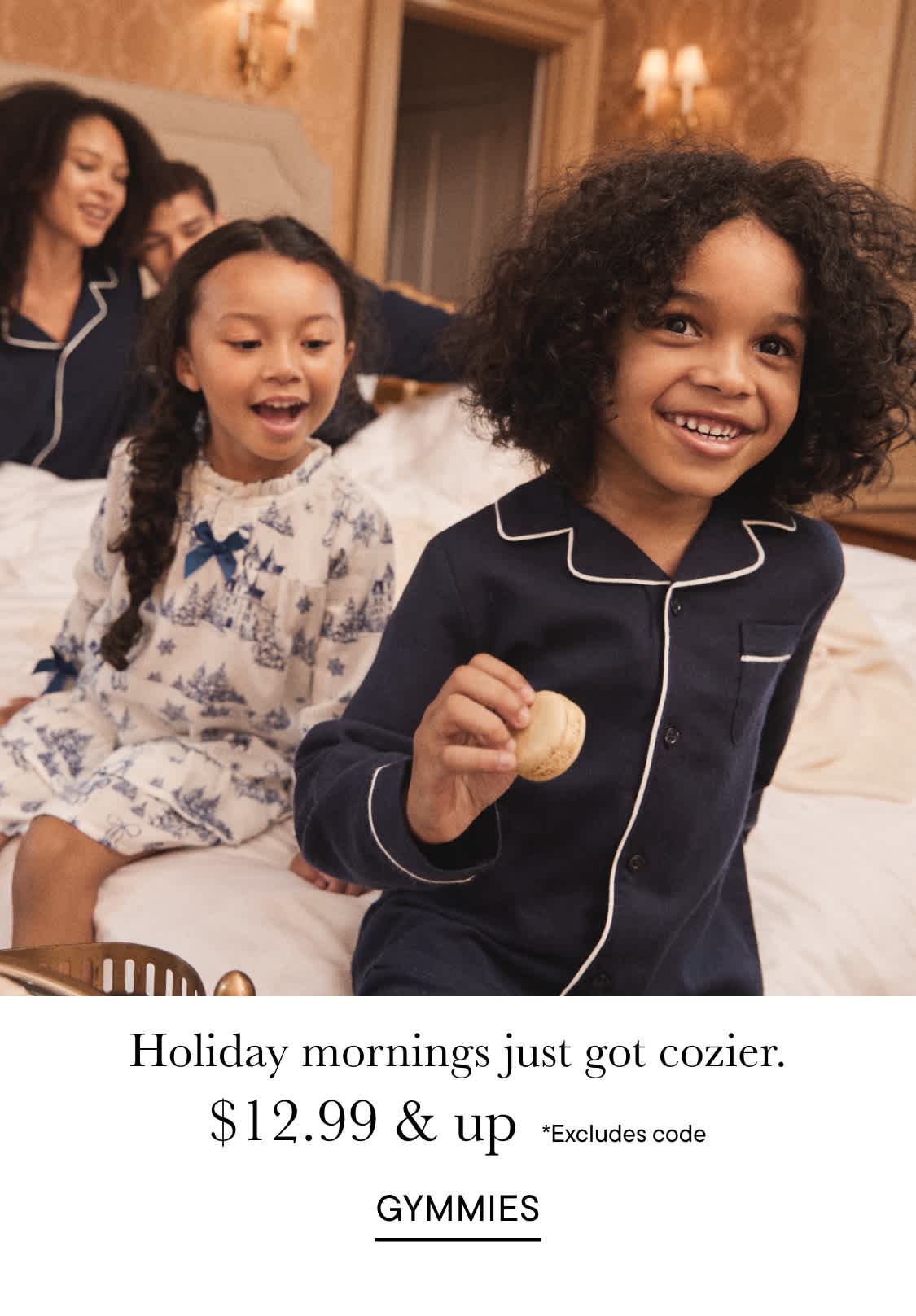 The image shows two young girls, one with curly hair and the other with straight hair, sitting on a couch and smiling. The background appears to be a cozy living room with a chandelier and other furnishings.