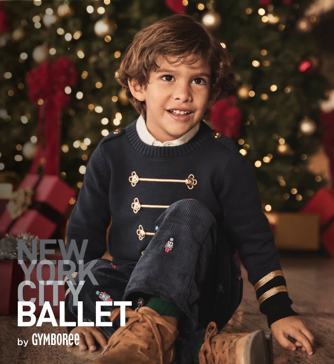 A young boy with curly hair is smiling in front of a Christmas tree with twinkling lights in the background.