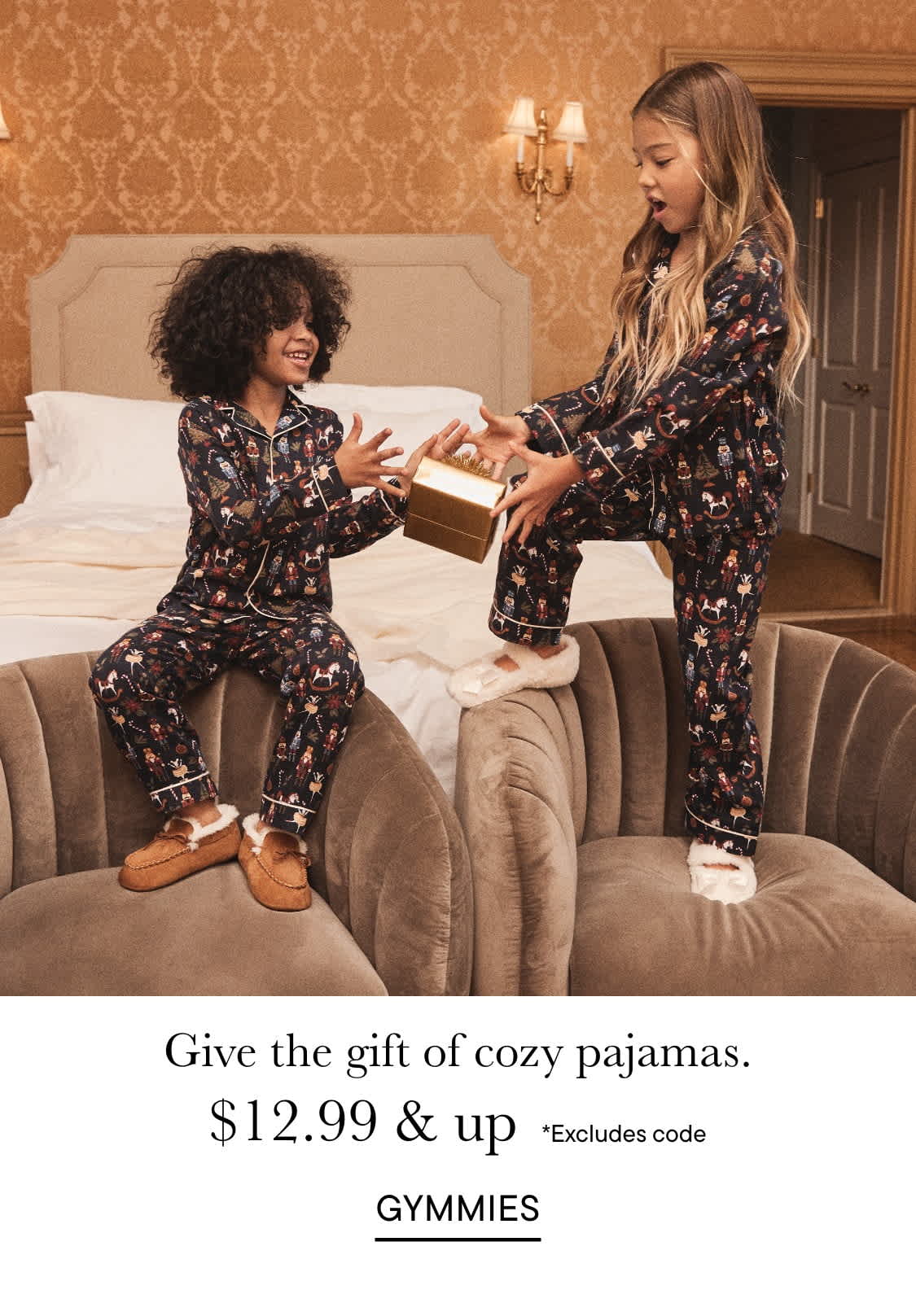 Two young women, one with curly hair and the other with long blonde hair, are sitting on a couch in a cozy-looking bedroom, wearing matching floral pajamas and smiling at each other as one of them holds a gift box.