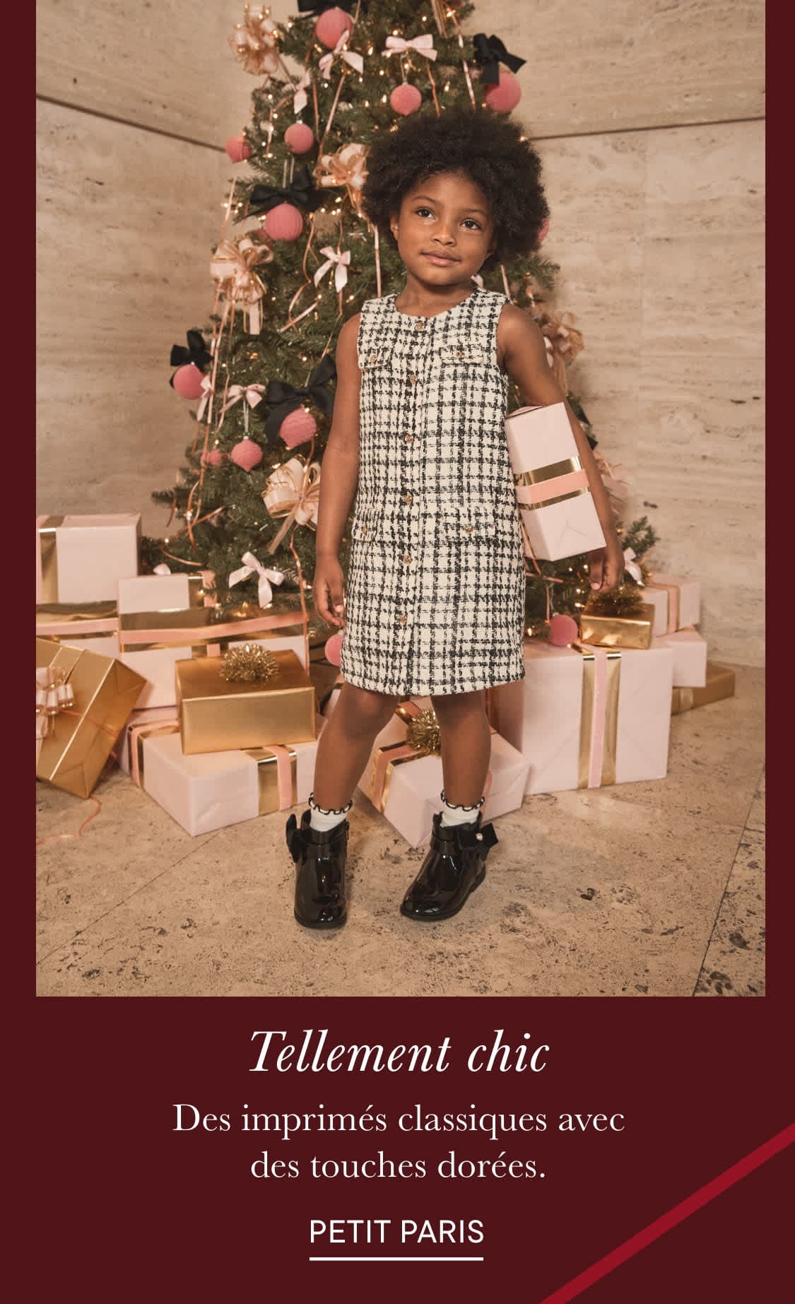 Une jeune enfant aux cheveux bouclés se tient devant un sapin de Noël décoré, vêtue d'une élégante robe à carreaux et de bottes noires, entourée de cadeaux emballés.