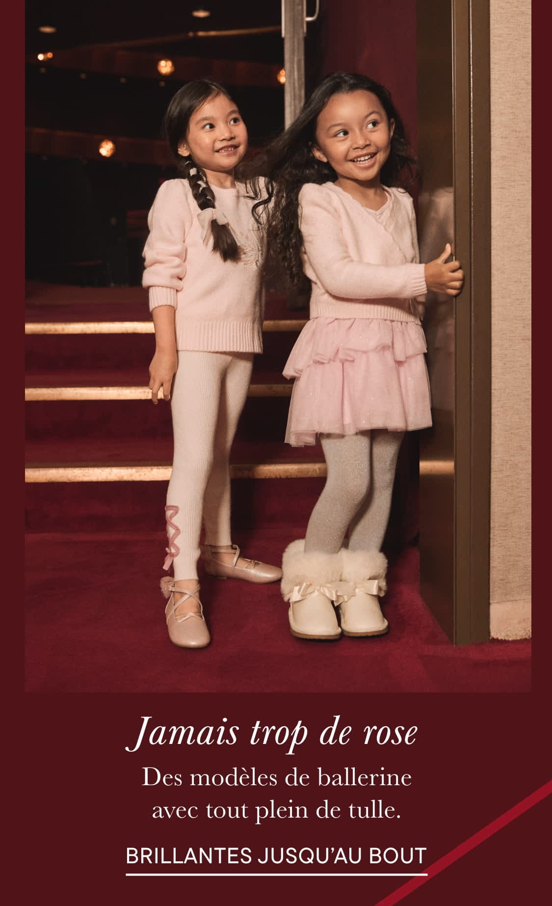 Deux jeunes filles vêtues de tutus et de pulls roses, debout sur un escalier recouvert de tapis rouge dans ce qui semble être un théâtre ou une salle de spectacle.