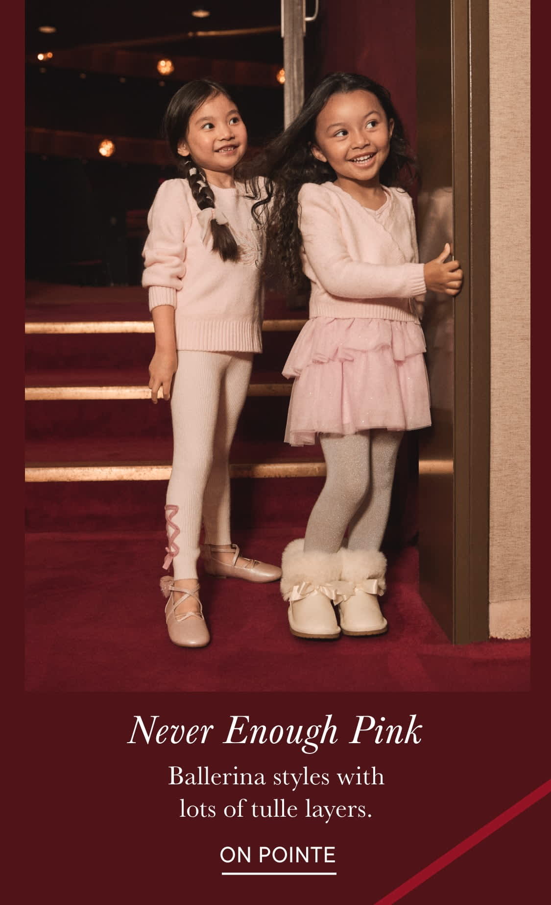 Two young girls dressed in pink tutus and sweaters, standing on a red-carpeted staircase in what appears to be a theater or performance venue.