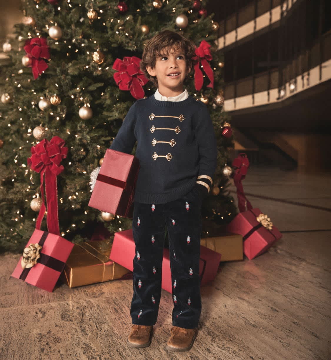 A young child wearing a navy blue sweater with gold buttons and polka dot pants stands in front of a Christmas tree decorated with red bows and ornaments, surrounded by wrapped gifts.