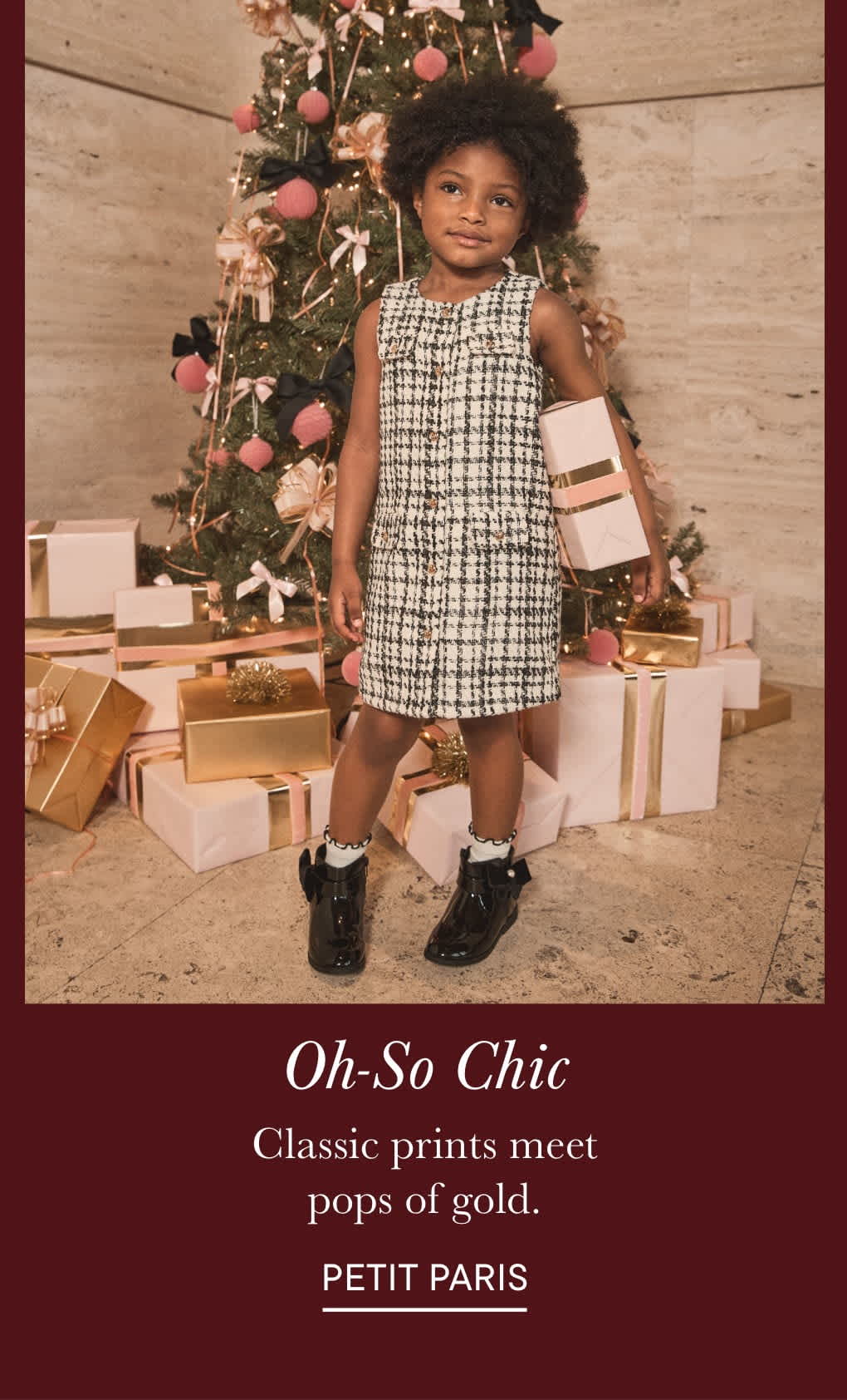A young child with curly hair stands in front of a Christmas tree, surrounded by wrapped gifts, wearing a checkered dress and black shoes.