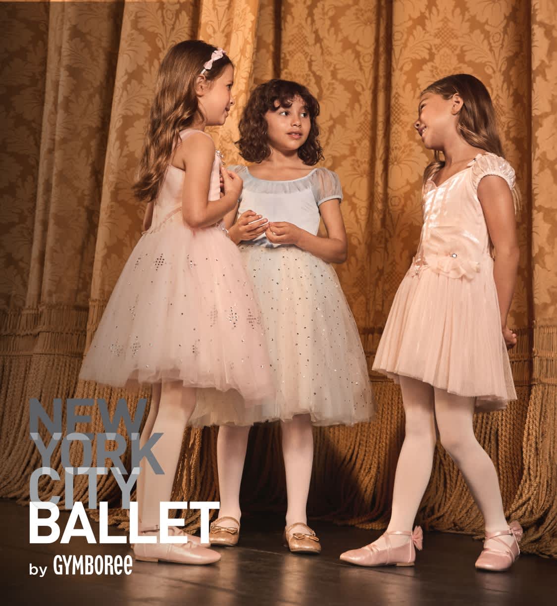 Three young girls in elegant, tulle-filled dresses stand together on a stage with a golden curtain backdrop.