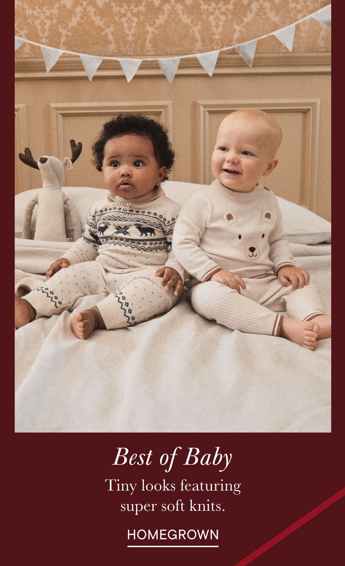 Two young children, a boy and a girl, sit together on a bed surrounded by decorative flags or banners. They are both wearing soft, patterned knit outfits and appear to be smiling and engaged with each other.