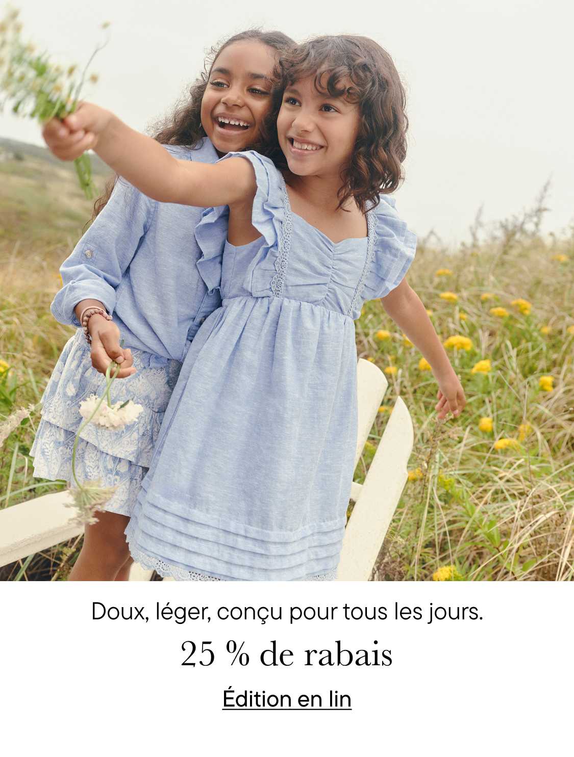 Deux jeunes filles en robes bleues s'enlacent et sourient joyeusement dans un champ de fleurs sauvages, avec une clôture en bois visible en arrière-plan.