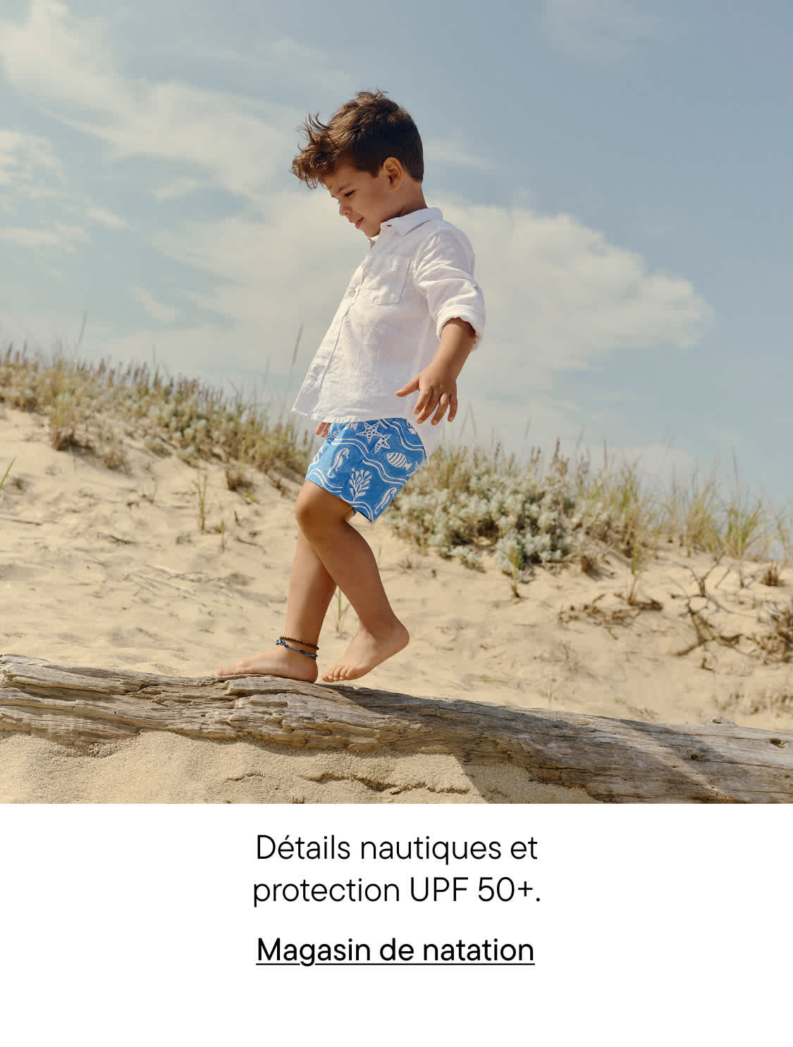 Un jeune enfant en chemise blanche et short de bain bleu à motifs marche le long d'un tronc d'arbre sur une plage de sable, avec un ciel nuageux en arrière-plan.