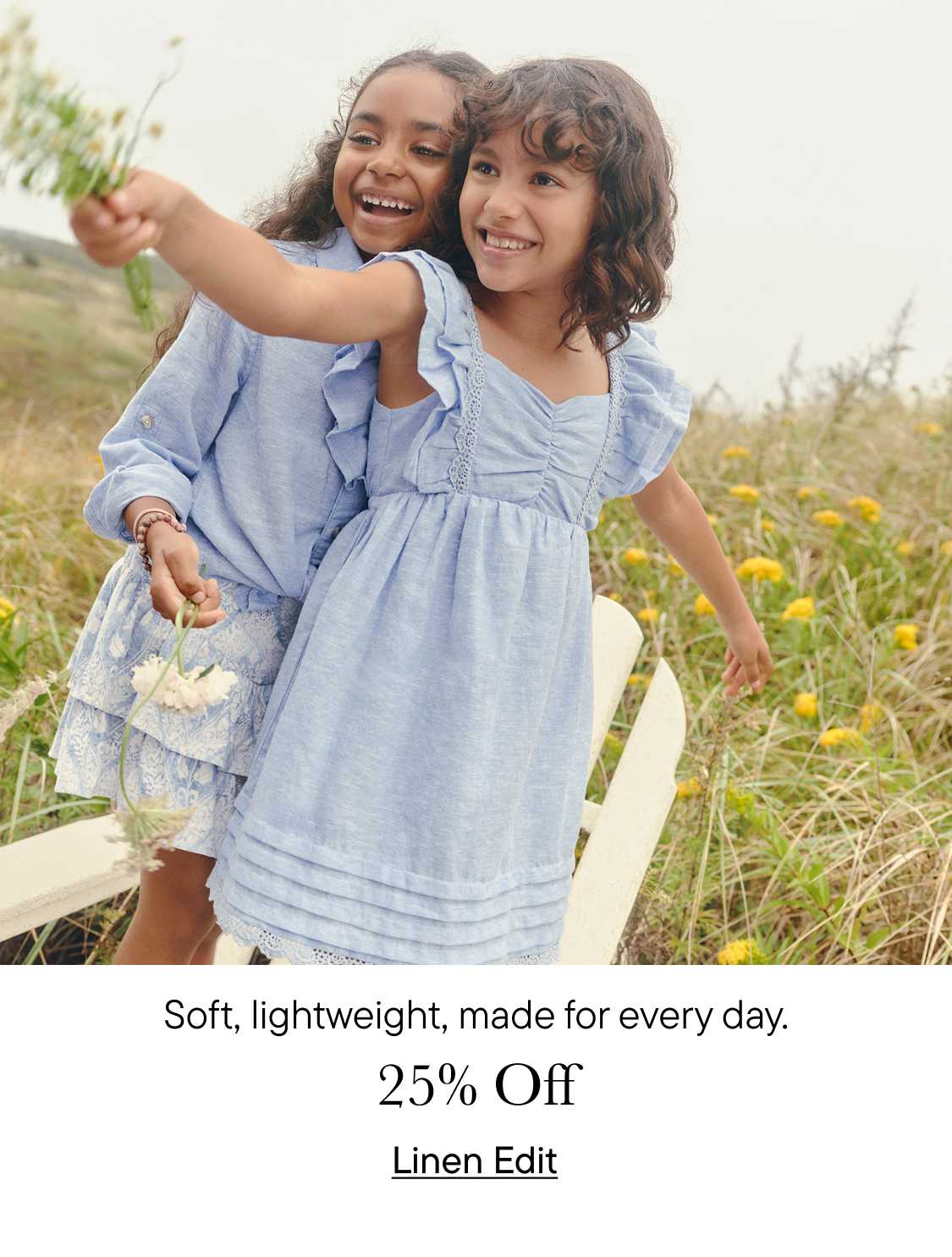 Two young girls in blue dresses are embracing and smiling joyfully in a field of wildflowers, with a wooden fence visible in the background.
