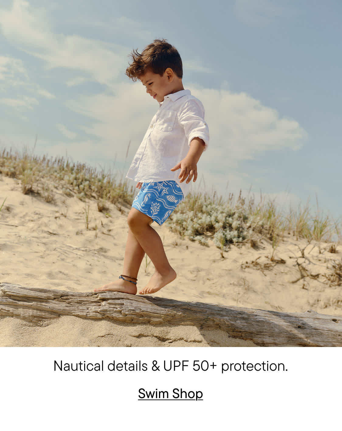 A young child in white shirt and blue patterned shorts walks along a wooden log on a sandy beach, with a cloudy sky in the background.