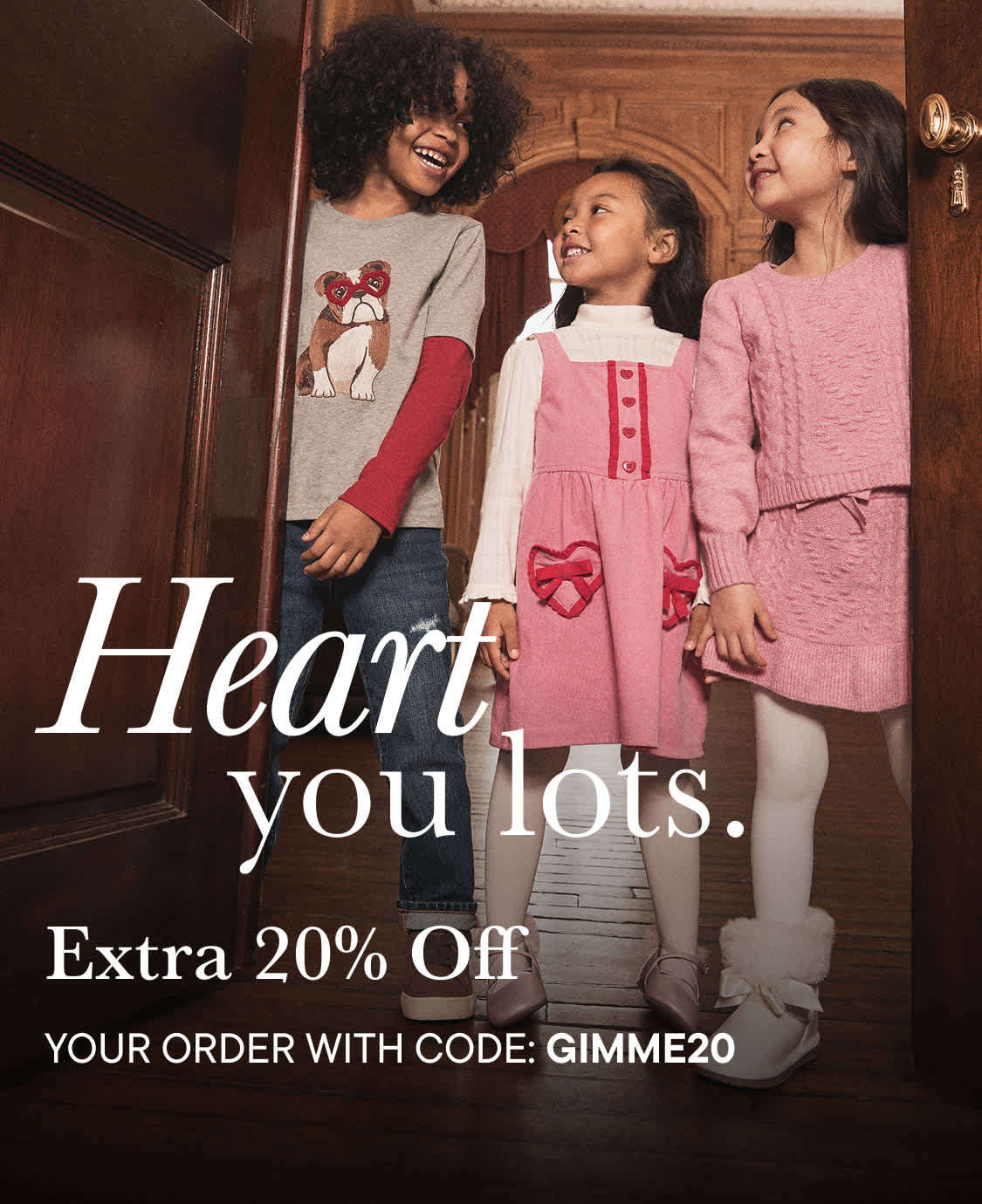 The image shows three young girls, two of whom are smiling and one who appears to be laughing, standing in front of a wooden bookshelf with a sign that says "I heart you lots." The girls are wearing casual clothing, including a gray sweater with a graphic design and a pink dress.