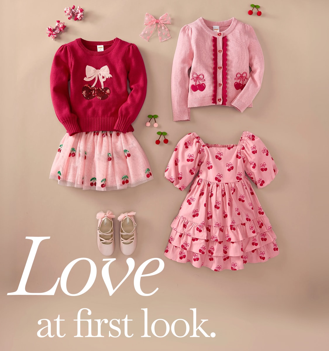 A collection of adorable pink and red children's clothing, including a sweater with a bow, a cardigan with cherry designs, a tulle skirt, and a patterned dress, all arranged on a light-colored background with the text "Love at first look" displayed.