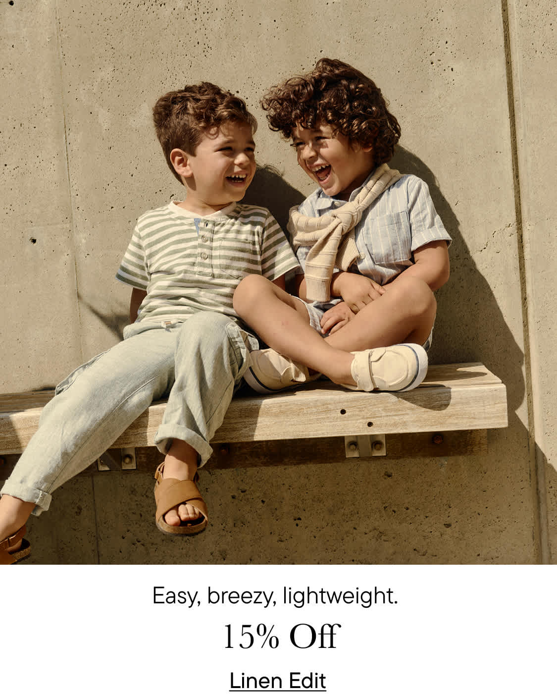 Two young boys, one with curly hair and the other with straight hair, are sitting together on a wooden bench against a plain background. They appear to be relaxed and comfortable with each other.