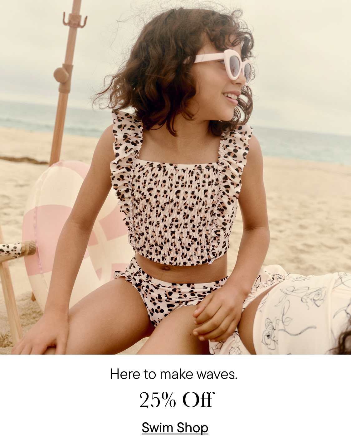 A young woman in a leopard-print swimsuit sits on a beach, smiling and ready to make waves, as indicated by the text "Here to make waves." in the background.