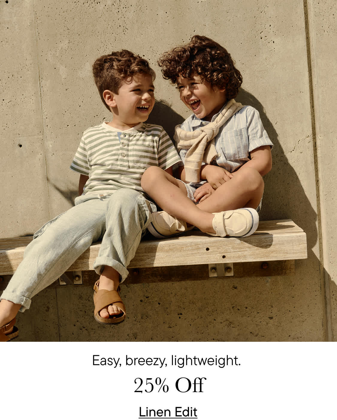 Two young boys, one with curly hair and the other with straight hair, are sitting together on a wooden bench against a plain background. They appear to be relaxed and comfortable with each other.