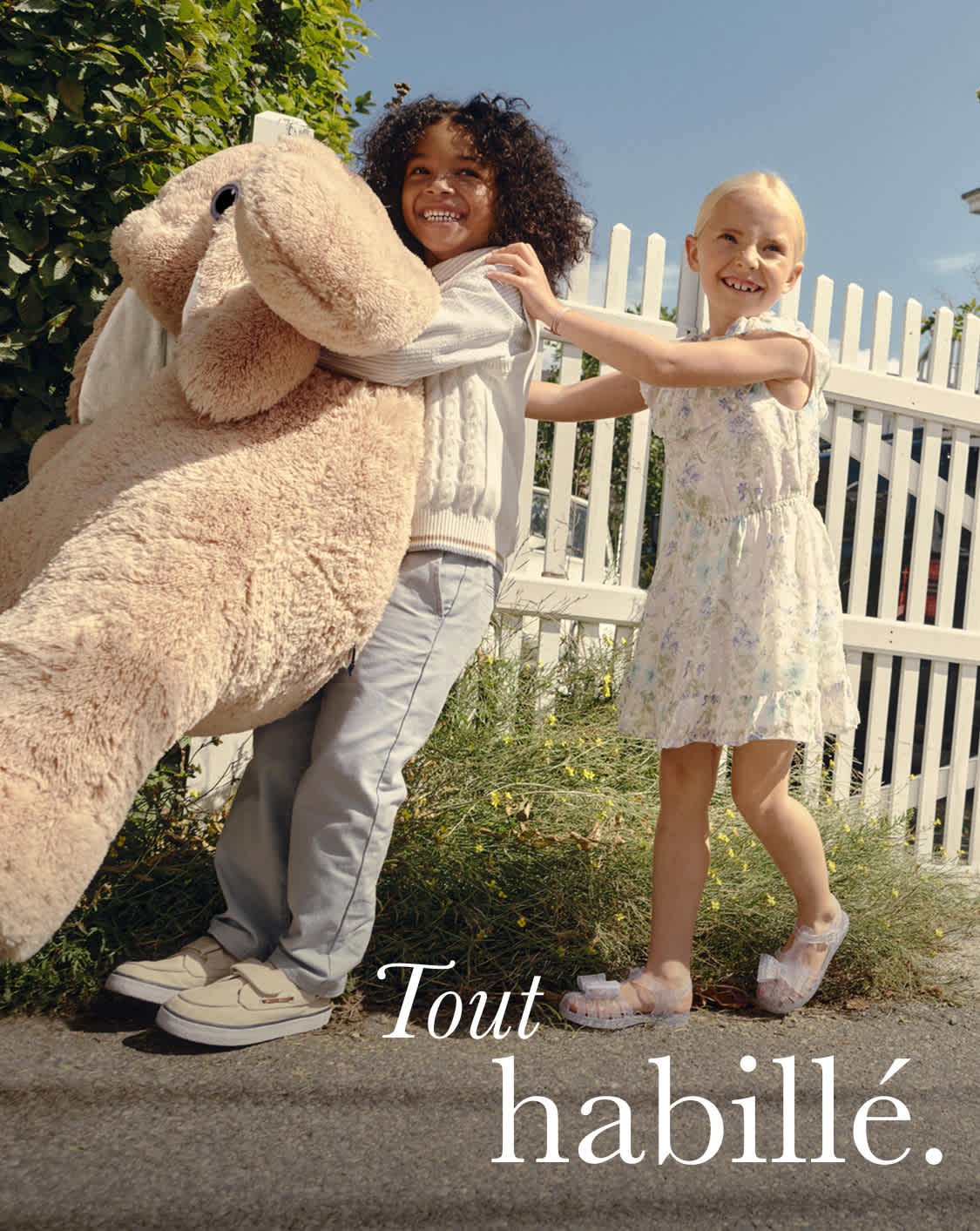 L'image représente un homme et une femme posant devant une clôture blanche entourée d'une végétation luxuriante. La femme porte un long manteau beige duveteux, tandis que l'homme est vêtu de façon décontractée. L'arrière-plan suggère un cadre extérieur, probablement un quartier résidentiel.