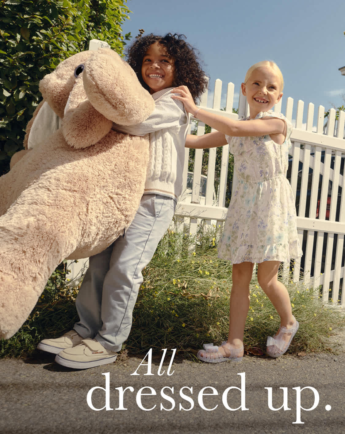 The image features two individuals, a man and a woman, posing in front of a white picket fence surrounded by lush greenery. The woman is wearing a long, fluffy beige coat, while the man is dressed in casual attire. The background suggests an outdoor setting, possibly a residential area.